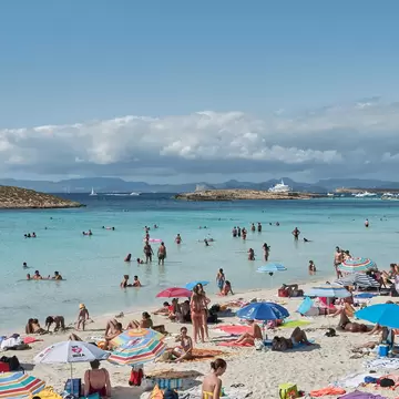 People under umbrellas relax on the sand of a large beach lapped by turquoise waves.