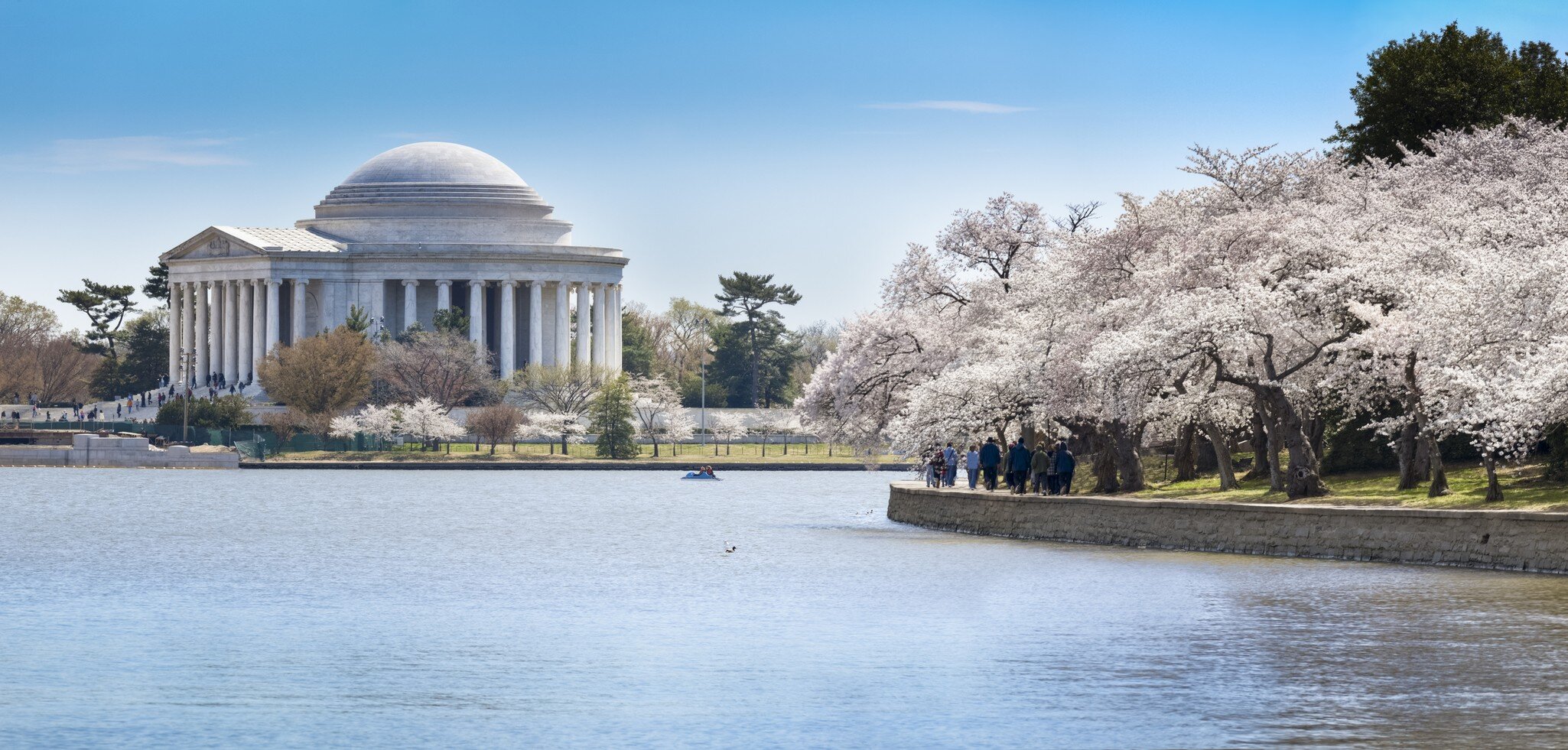 Domed marble rotunda ringed by columns across calm water; walkers on a stone seawall and a small boat mid-basin.