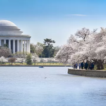 Domed marble rotunda ringed by columns across calm water; walkers on a stone seawall and a small boat mid-basin.