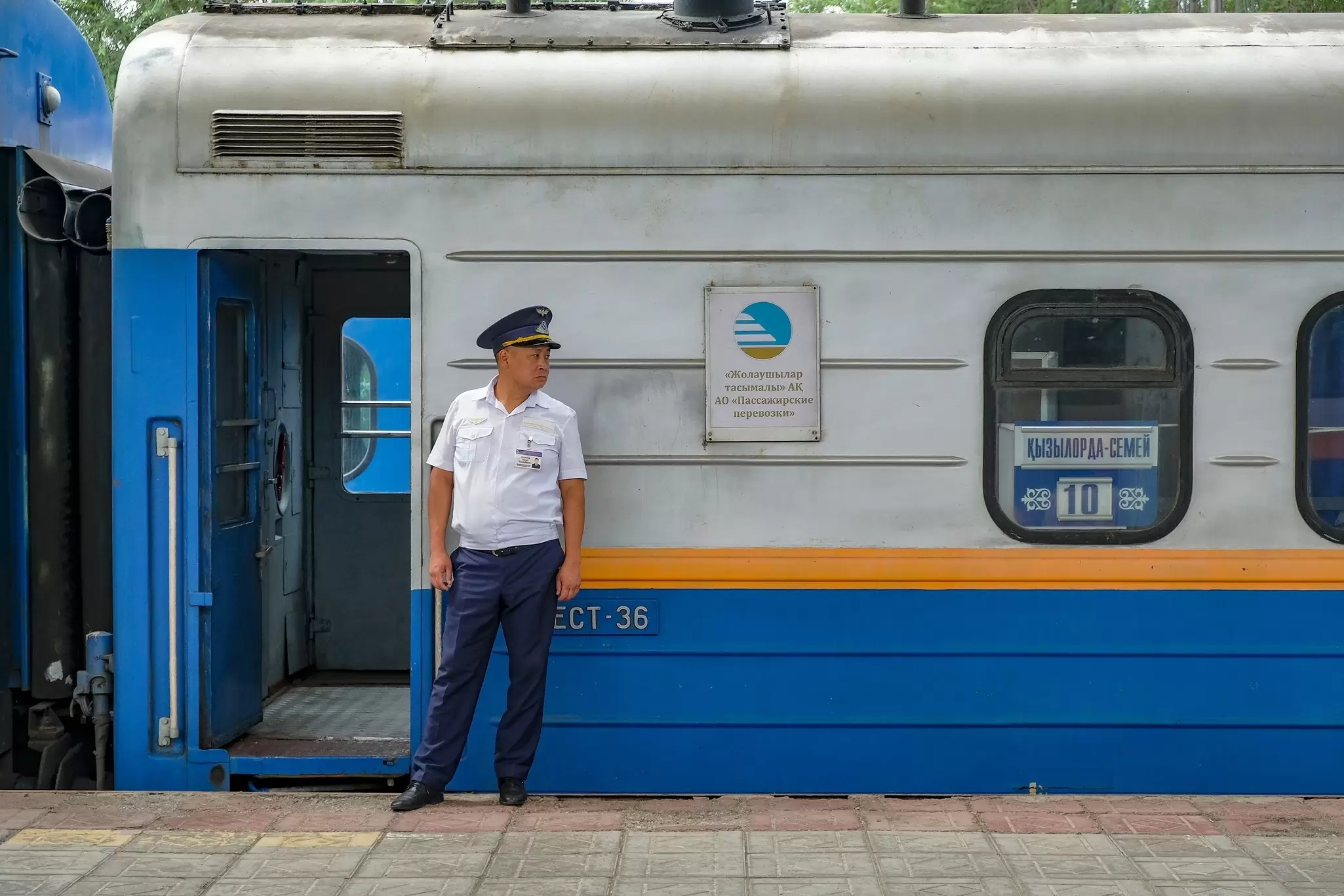 An inspector in a unform stands in front a train car with blue and yellow livery in the station at Alatau (formerly Zhetygen), Kazakhstan