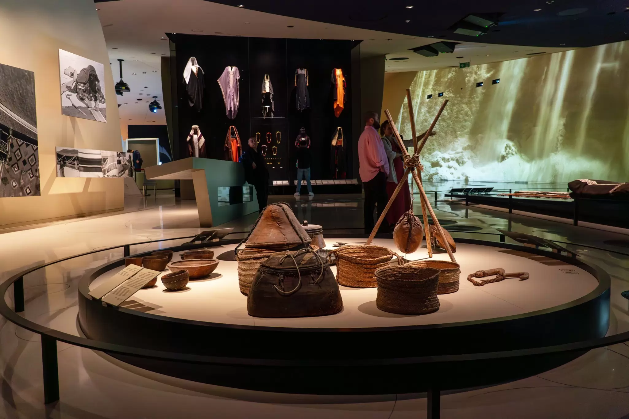 Woven baskets and traditional objects on a circular platform in a dim museum gallery with costumes in a glass case behind.