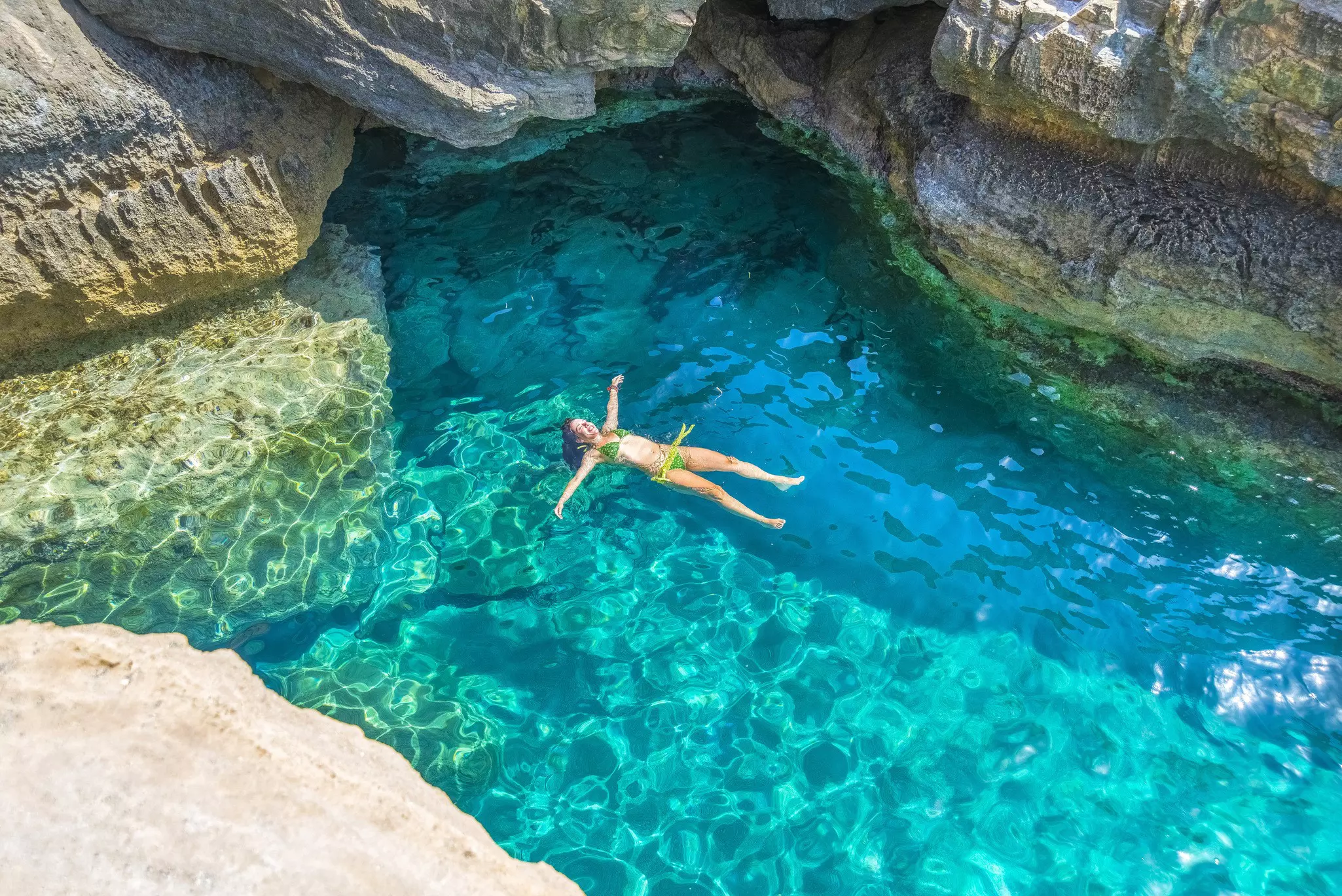 A view from above of a woman in a bikini floating in blue waters in a rocky cove.