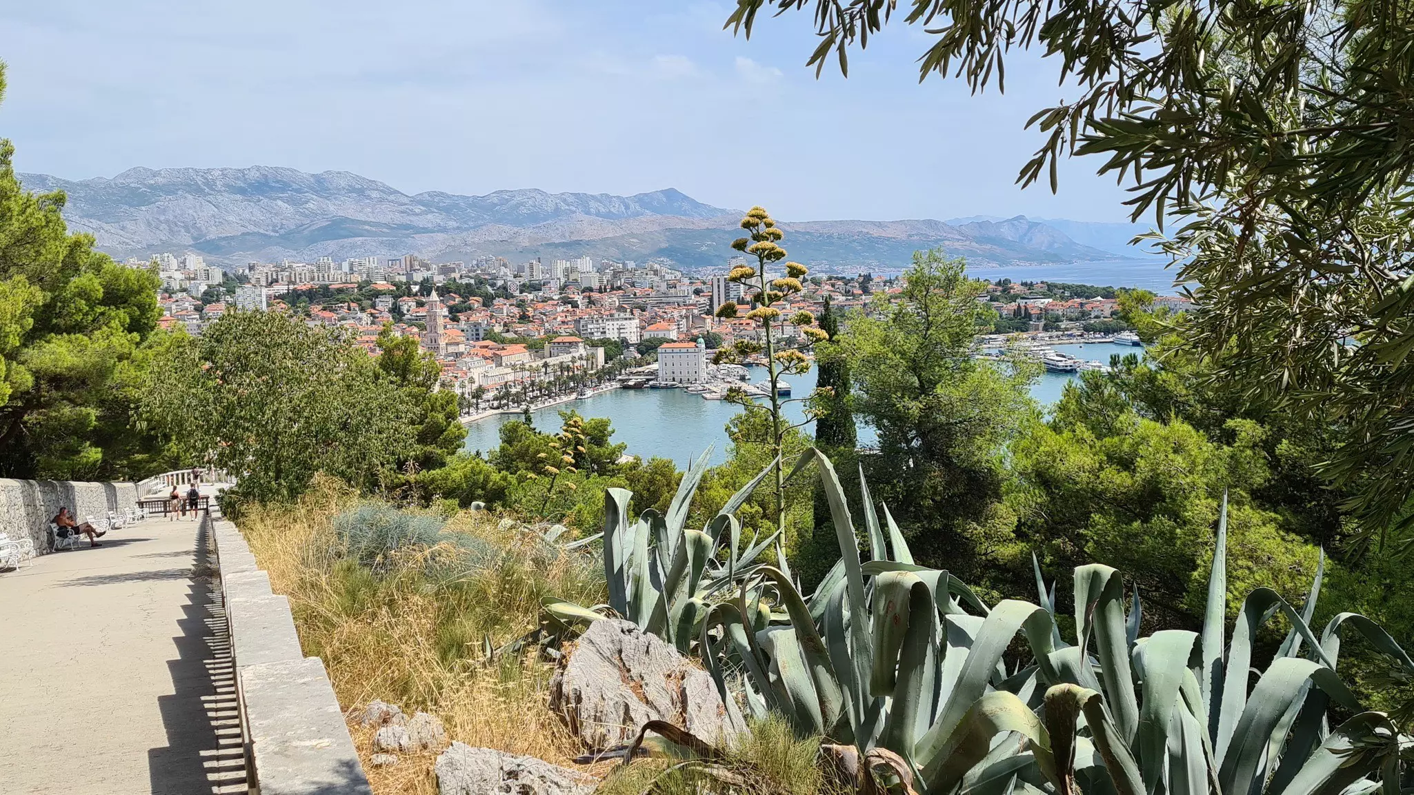 A long steep path leads up a hill with views back towards a coastal town with red-roofed buildings and a small harbor.