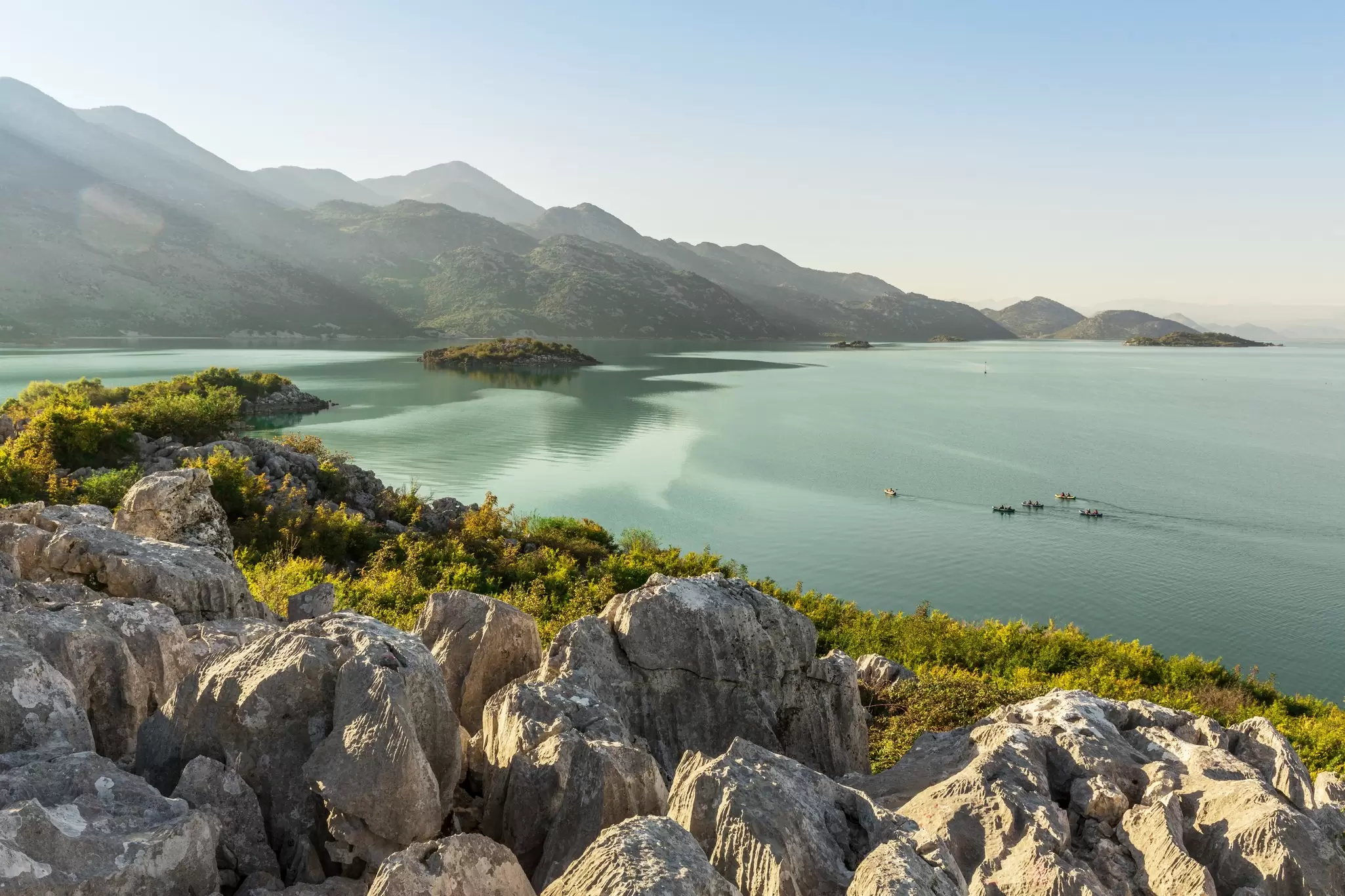 Kayakers make their way across a vast lake surrounded by craggy hills with greenery