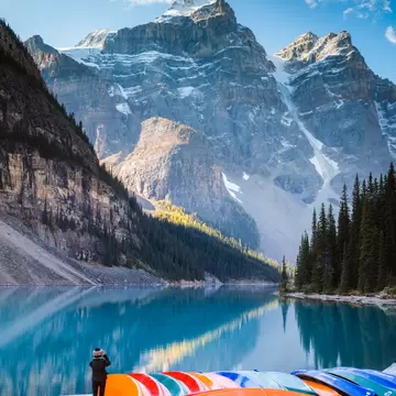 Woman taking a photo of Moraine Lake, in Banff National Park, next to a stack of colorful canoes in autumn.