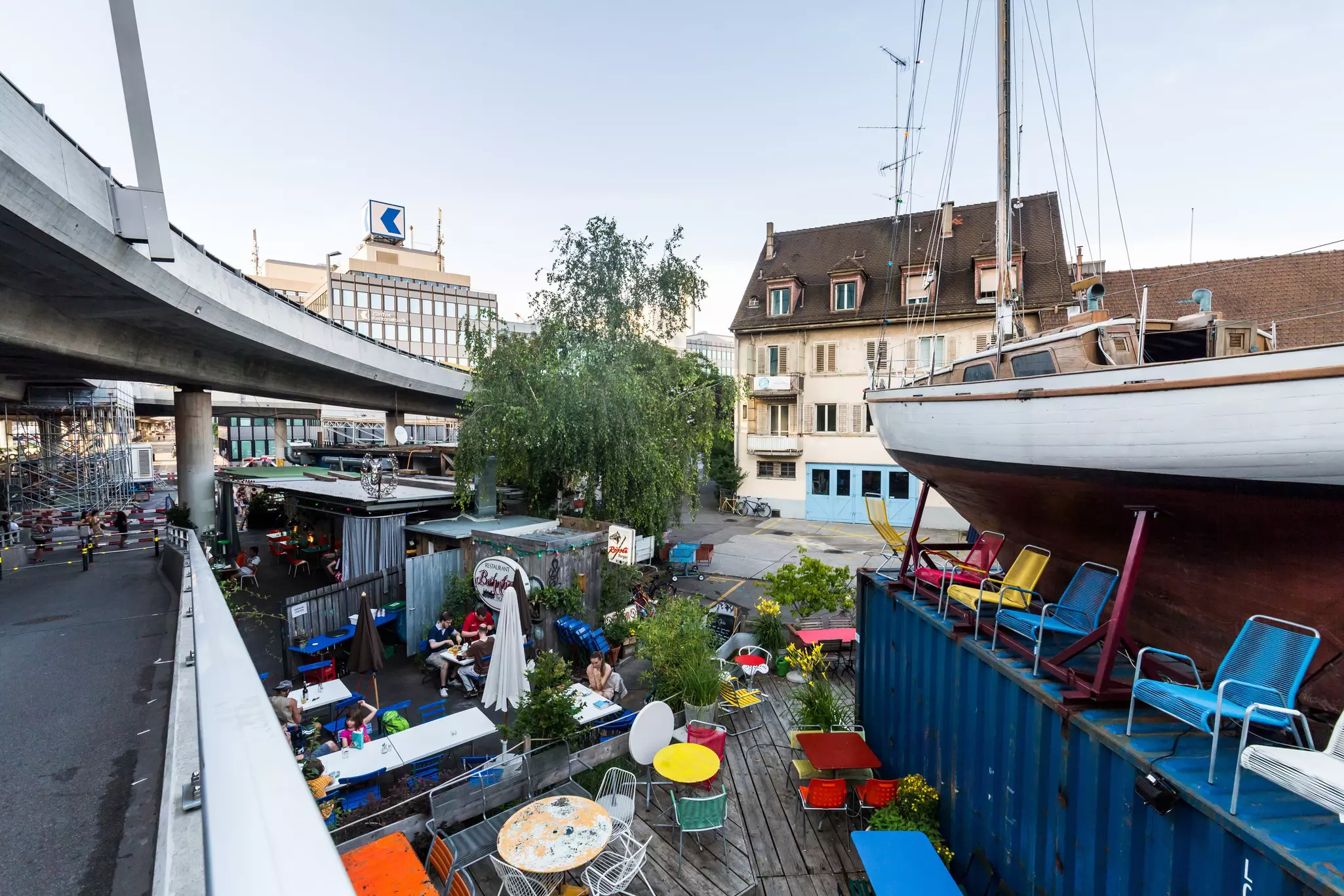 A cafe with colorful chairs occupies a space in a city neighborhood next to a boat and shipping container, and highway overpass.