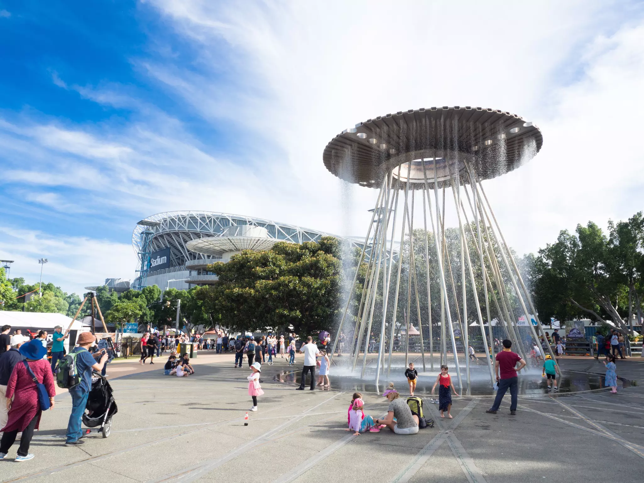 Families gather around a large fountain with circular top in the middle of a public sqaure on a sunny day.