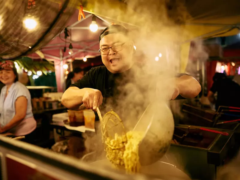 A food stall operator stirs a pot of asian food as billowing steam fills the air