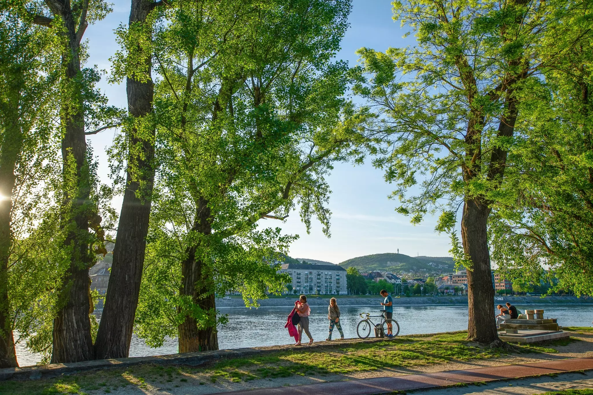 Two people walking by the riverbank in Budapest; another person is standing with a bicycle, and a couple is sitting on a bench.