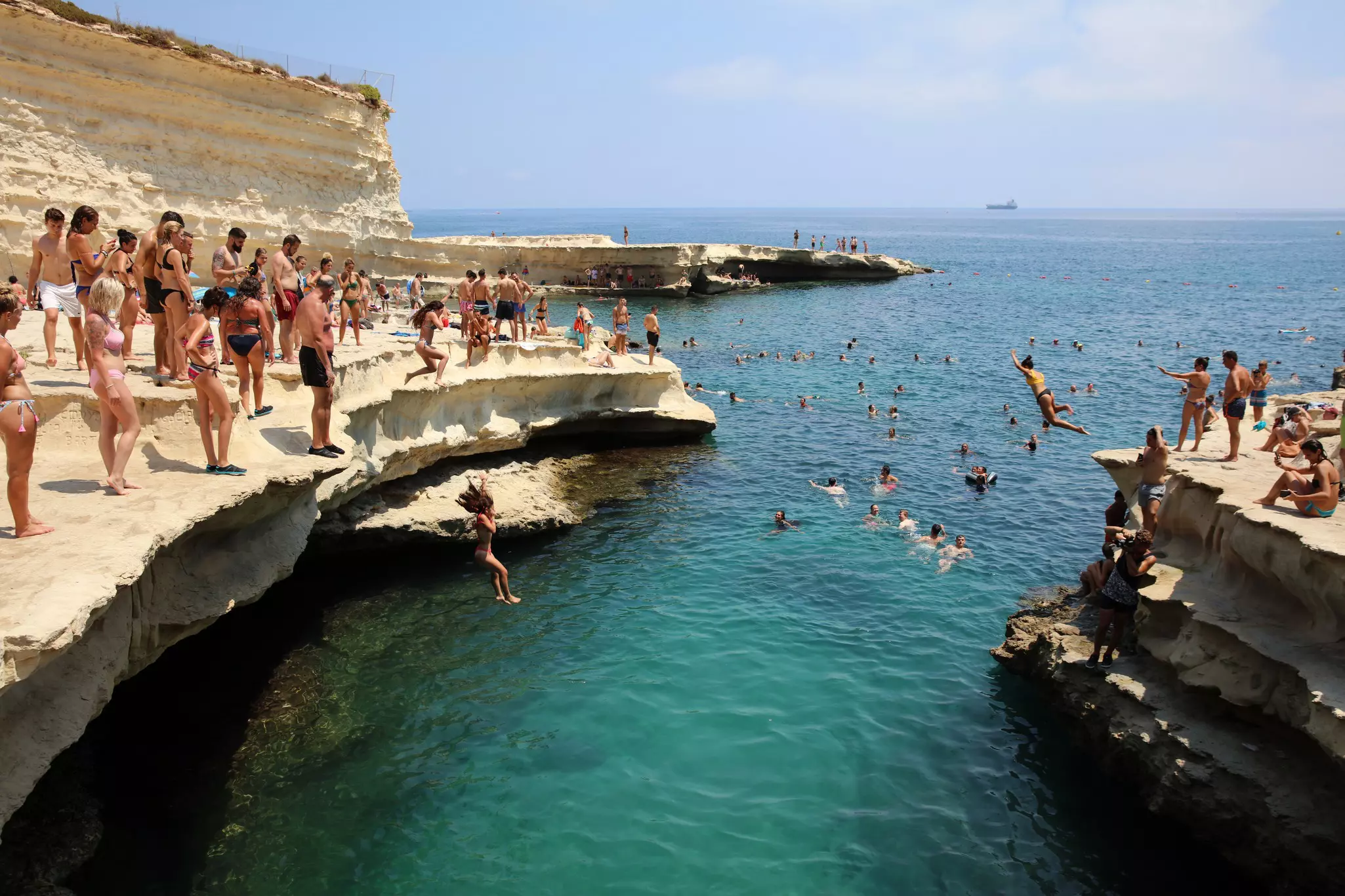 People in swimsuits stand on a rocky ledge and watch people jump into the blue ocean below.