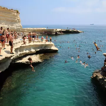 People in swimsuits stand on a rocky ledge and watch people jump into the blue ocean below.