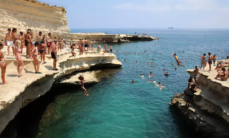 People in swimsuits stand on a rocky ledge and watch people jump into the blue ocean below.