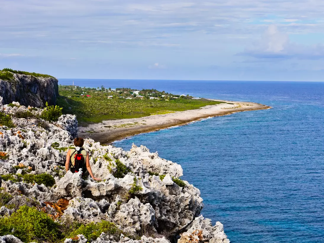 Hiker on jagged limestone at the Bluff, Cayman Brac, looking along a shore of beach and scrub toward the open sea