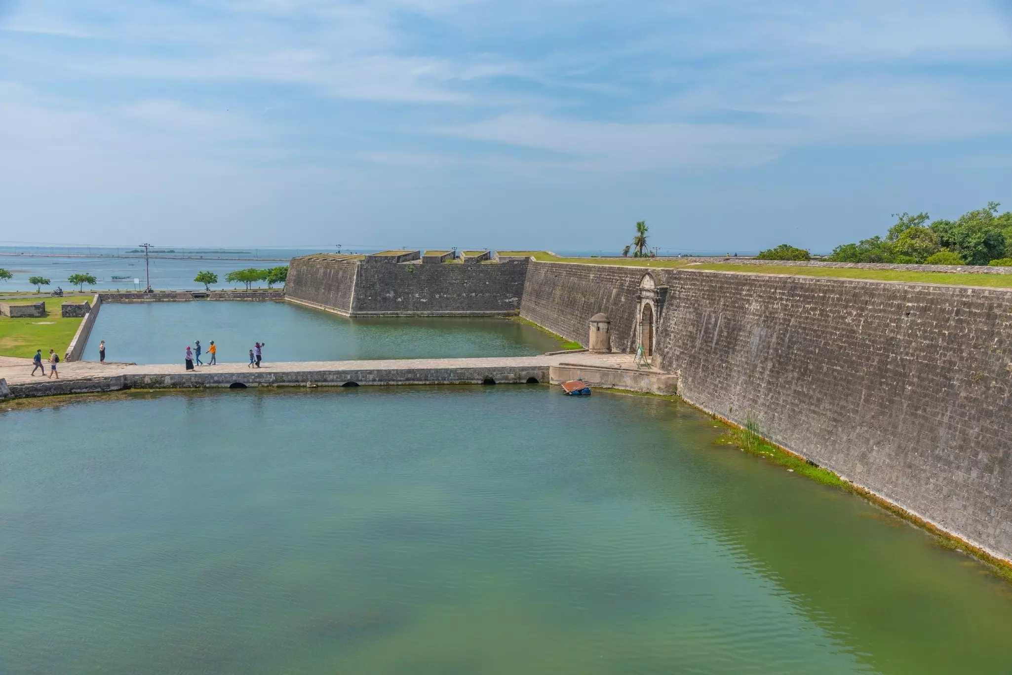 People walk across a bridge over a moat leading to a large coastal fortress.