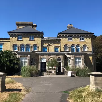 A yellow-brick Italianate Victorian villa with two hipped-roof wings, arched windows and a driveway under a clear blue sky