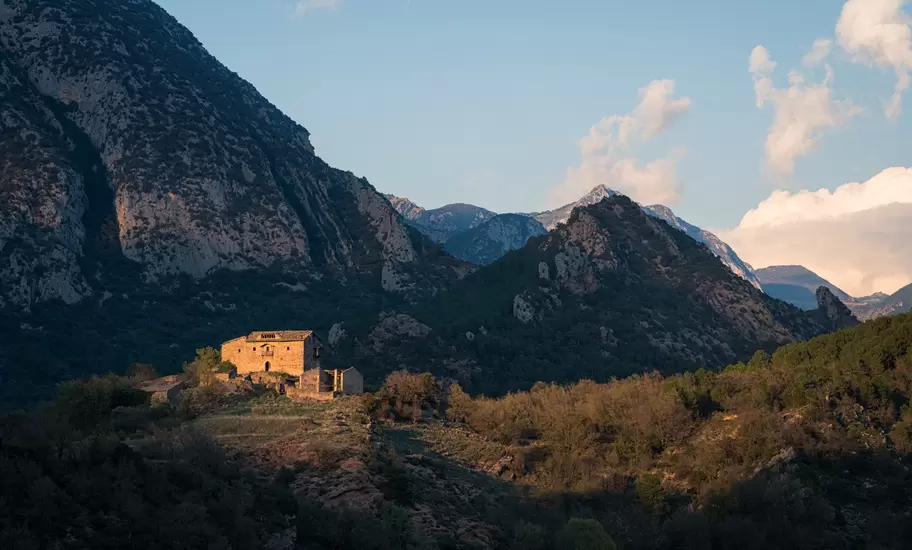 Stone farmhouse lit by low sun on a wooded slope beneath rocky mountain peaks in the Andorran Pyrenees