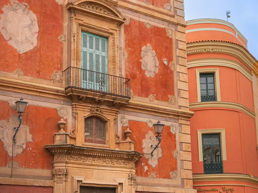A rococo palace front in salmon and ochre with a carved stone portal and wrought-iron balconies, people crossing the square in front.