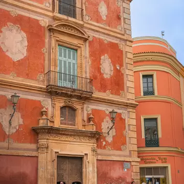 A rococo palace front in salmon and ochre with a carved stone portal and wrought-iron balconies, people crossing the square in front.