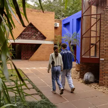 Two people walking along a tiled path between red-brick buildings and a vivid blue wall, framed by leaves.