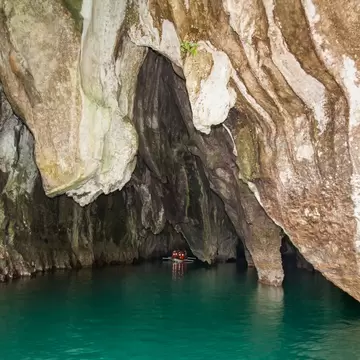 Massive streaked limestone cave walls overhanging green water, with a small boat of visitors in red life jackets far inside.