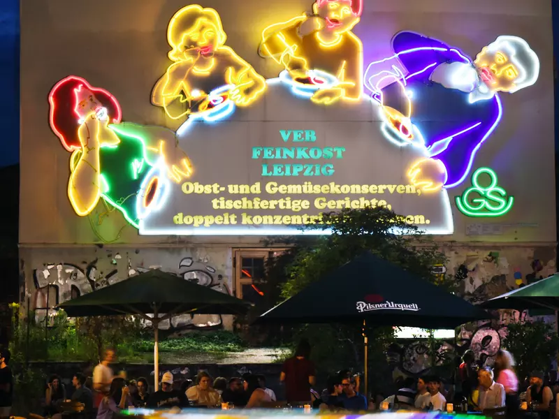 Patrons at a Leipzig beer garden sit at tables underneath a neon sign showing a family eating with spoons