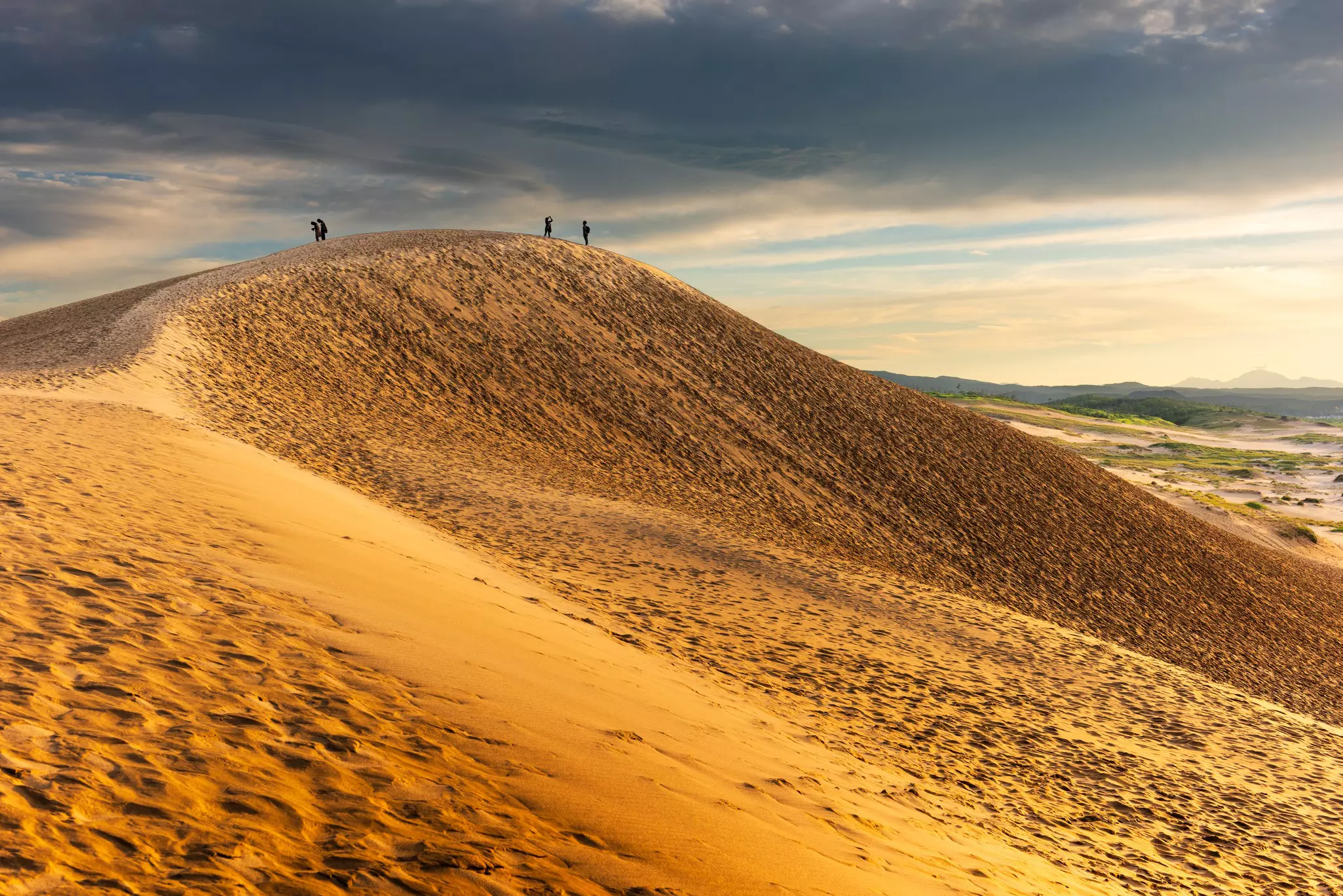 Four people atop large sand dunes with a cloudy sky in the background