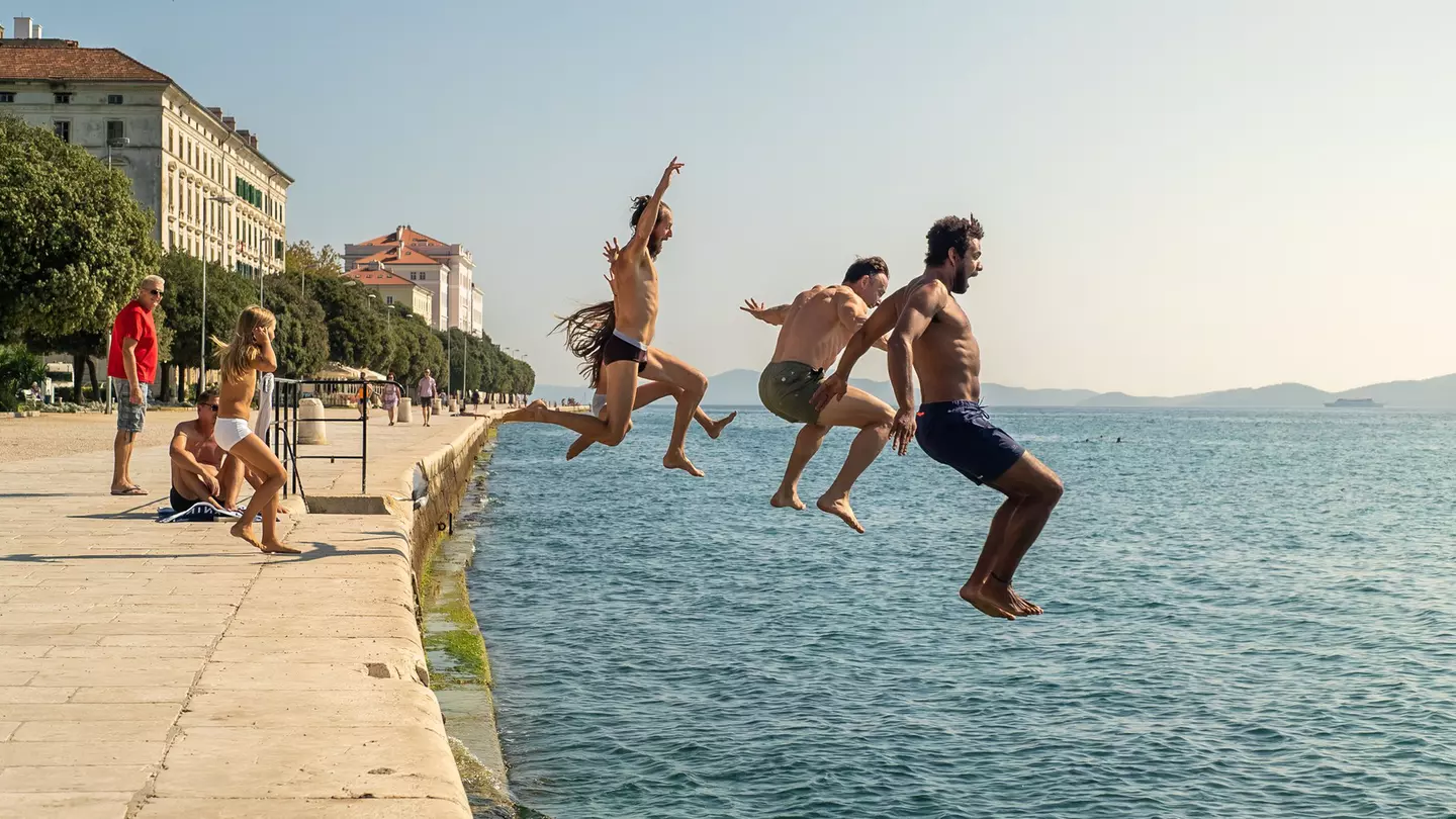A group of people are pictured midair as they jump off the harbor of a town into the sea.