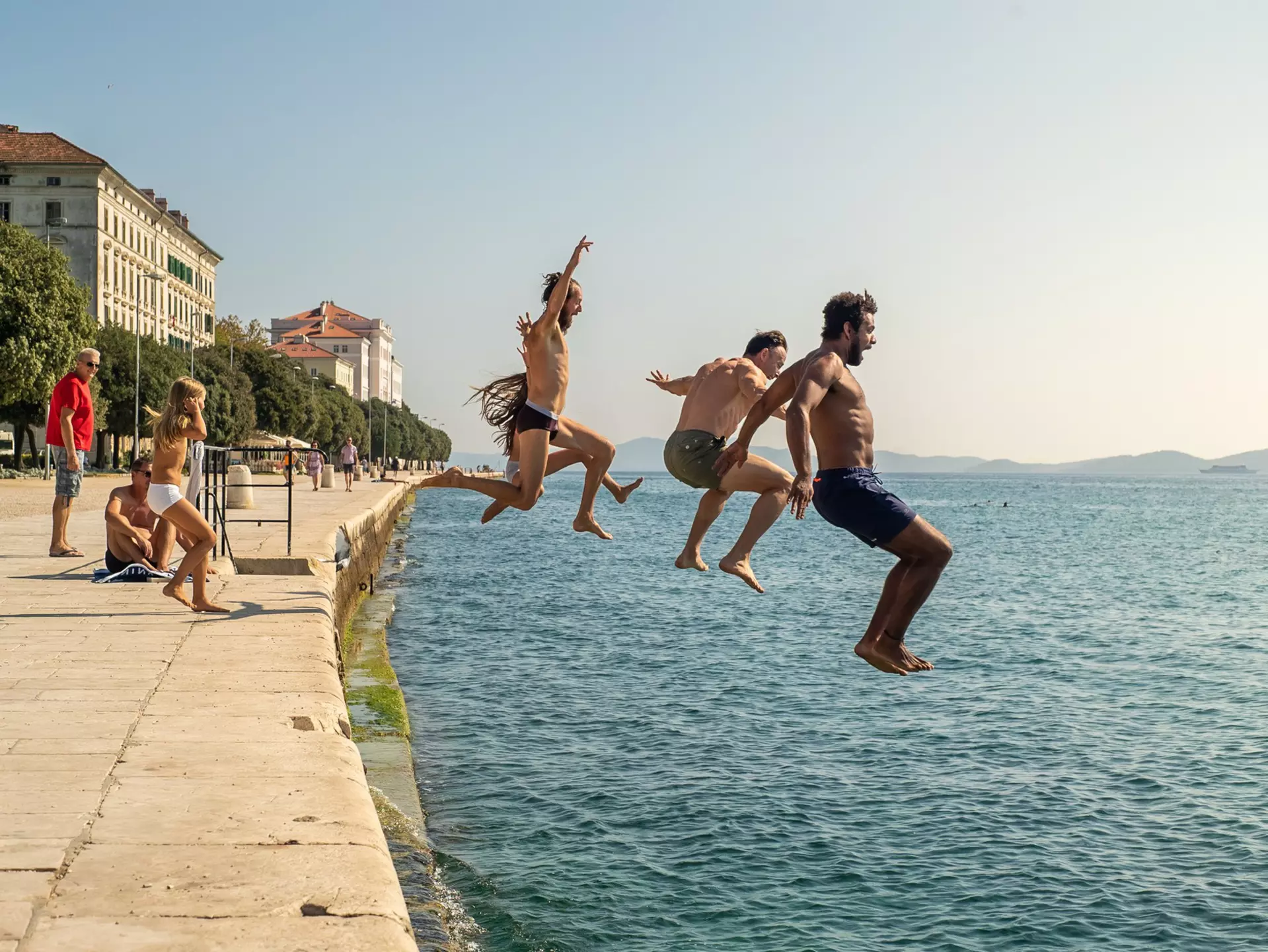 A group of people are pictured midair as they jump off the harbor of a town into the sea.