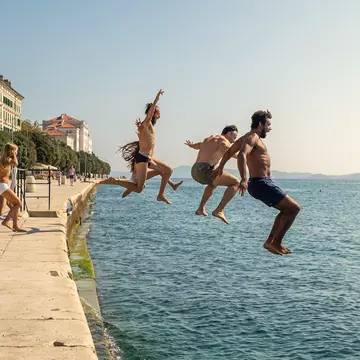 A group of people are pictured midair as they jump off the harbor of a town into the sea.