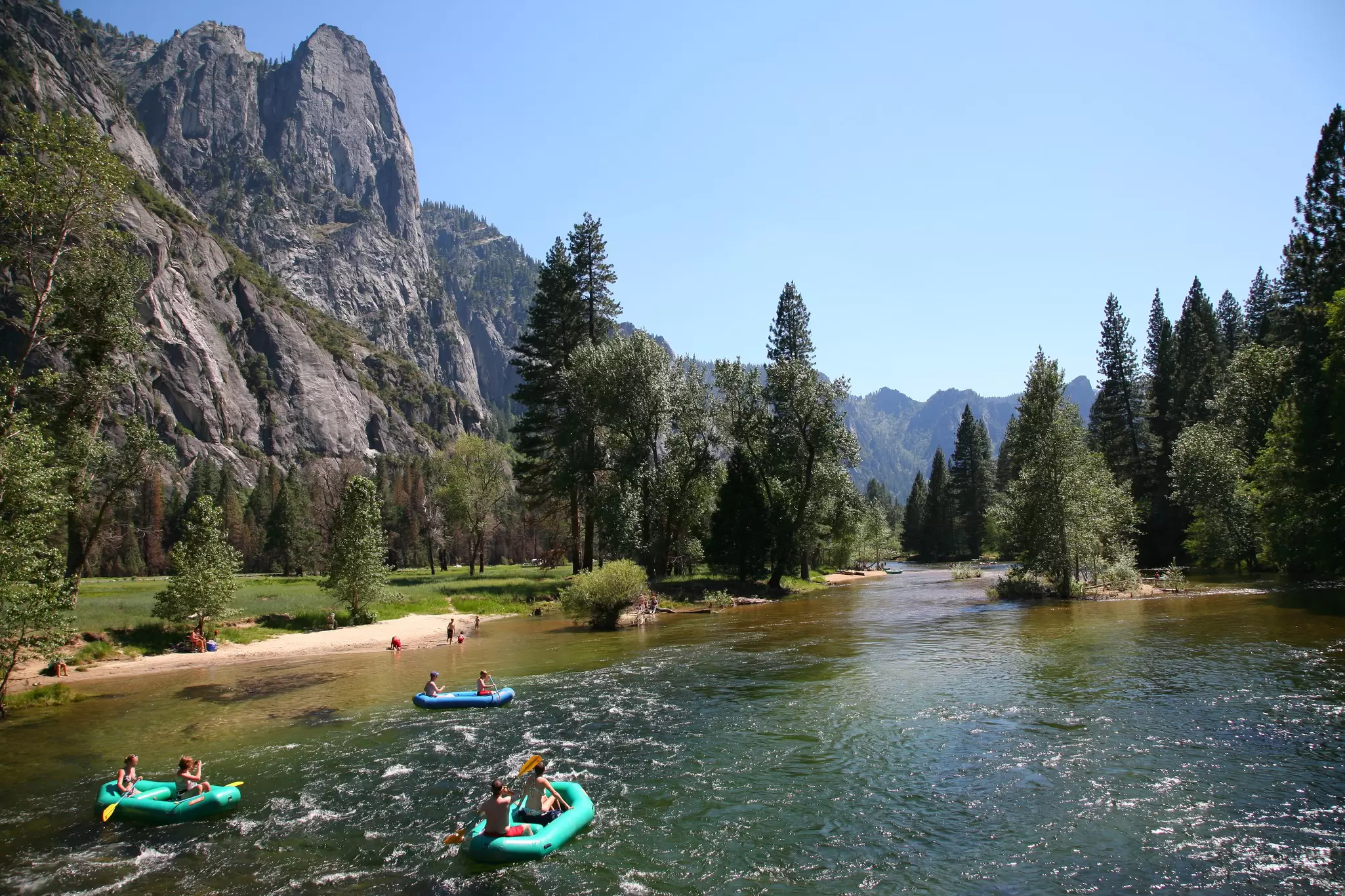 People rafting in a river surrounded by pine trees and large rocky mountain. 