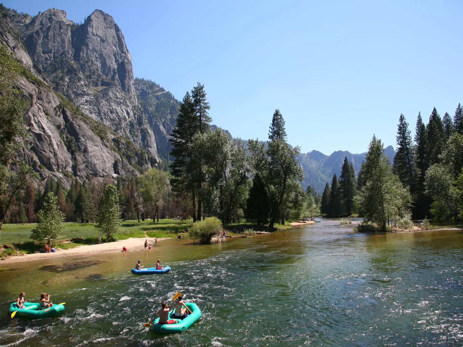 People rafting in a river surrounded by pine trees and large rocky mountain. 