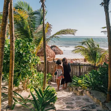 2 girls walking towards the ocean on a path surrounded by palm trees and greenery.