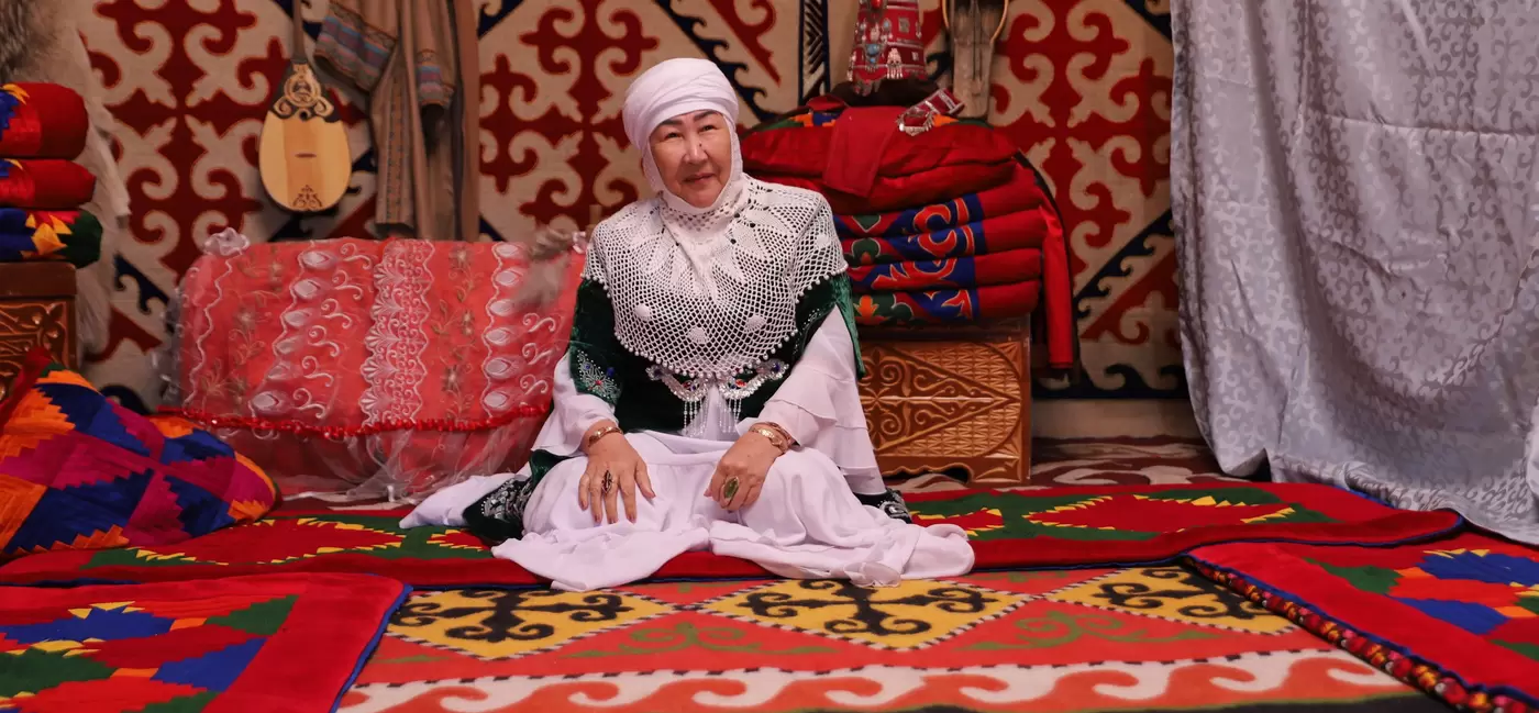 A woman in traditional Kazakh dress sits in a room draped with red patterned carpets and furnishings