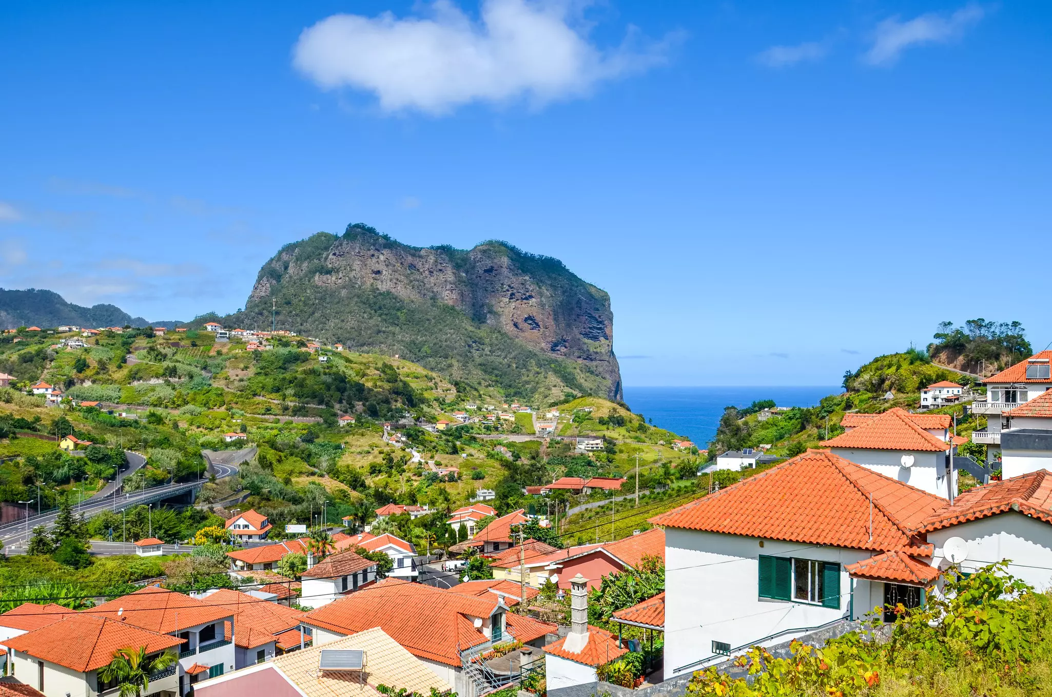 A village high up on a hill tucked around a massive cliff-top boulder, with views down towards the distant ocean. 