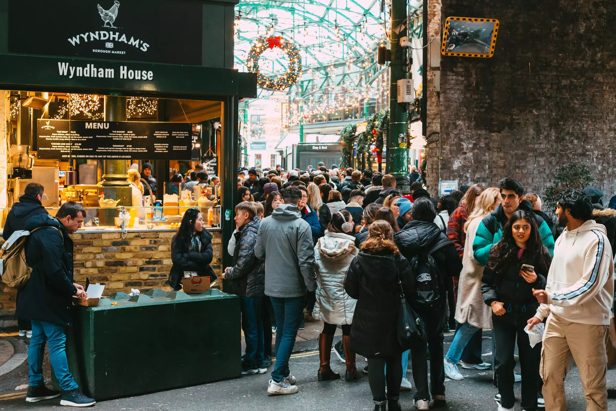 Shoppers pass by a food stall in a vast covered market that's decorated for the festive season, with a large wreath hanging from above and string lights between stalls.