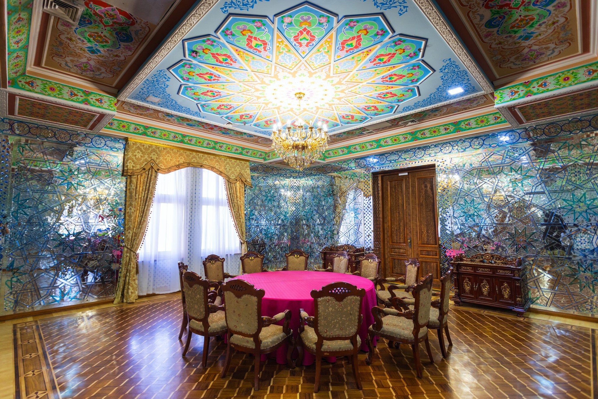 A mirrored reception room with a hand-painted domed ceiling in blue, green and red, a gilt chandelier above a round table laid with pink cloth and carved chairs.