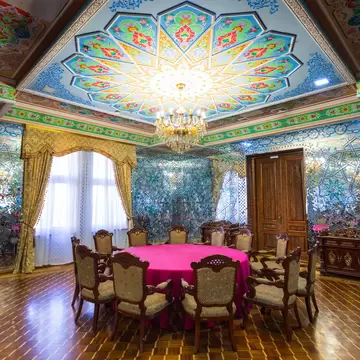 A mirrored reception room with a hand-painted domed ceiling in blue, green and red, a gilt chandelier above a round table laid with pink cloth and carved chairs.