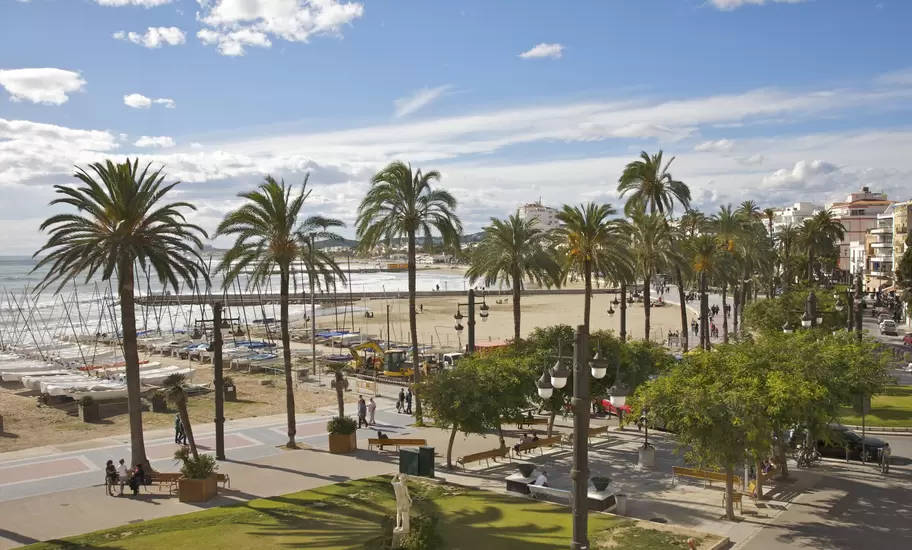 Sitges, Spain: palm-lined seafront promenade with benches and a white statue, sailing dinghies drawn up on the beach