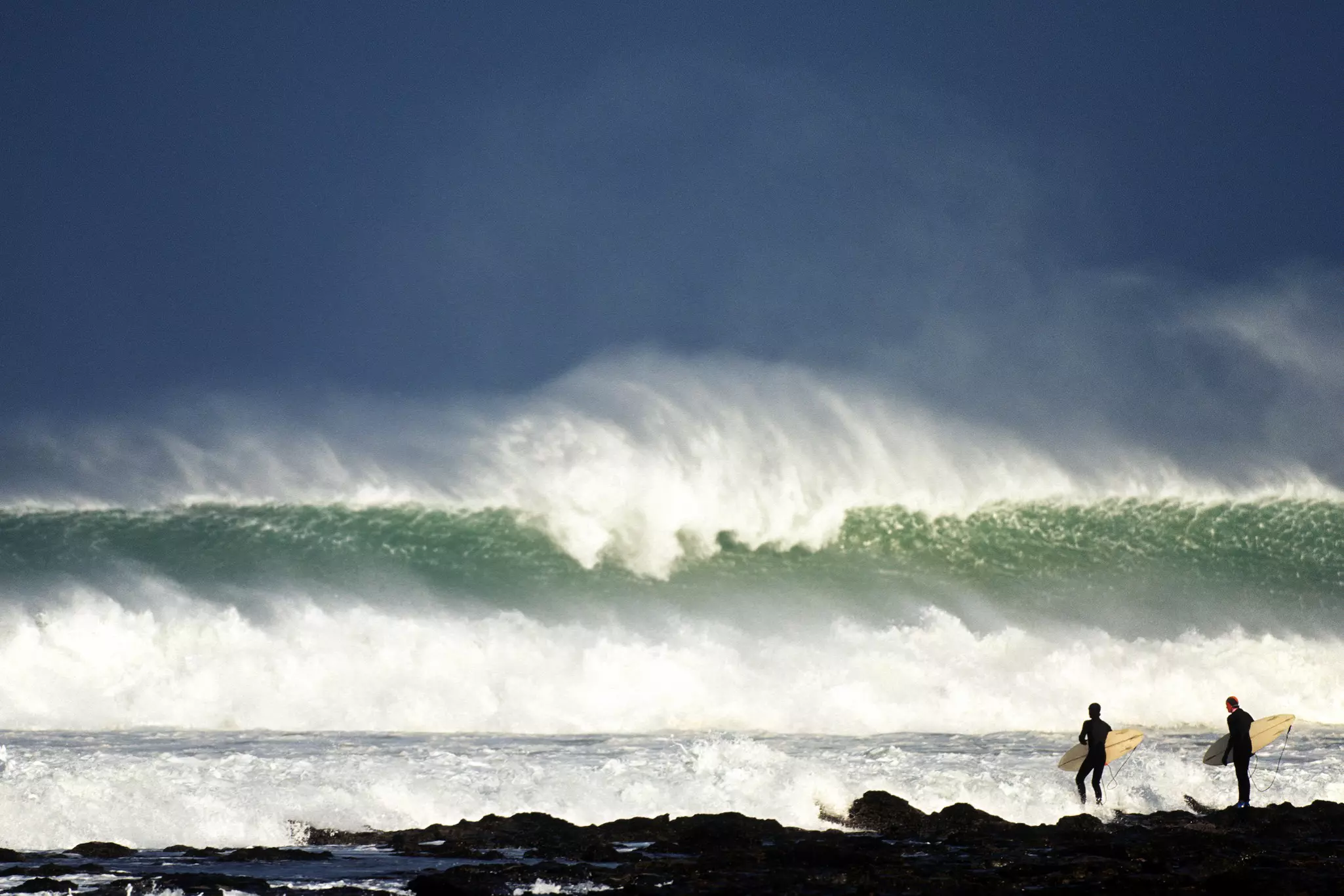 Two surfers in full wet suits stand with their boards on the beach looking out towards the huge waves crashing on the break.