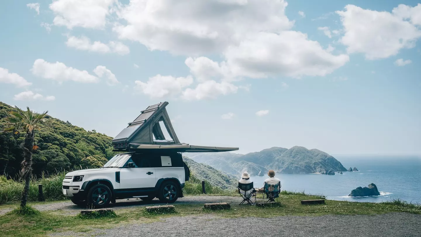 Couple sitting in a camping chair in nature with a view of Island in a beautiful blue sky with an overlanding car with a rooftop tent and an awning. Hokkaido 1, other: Kat Marsh