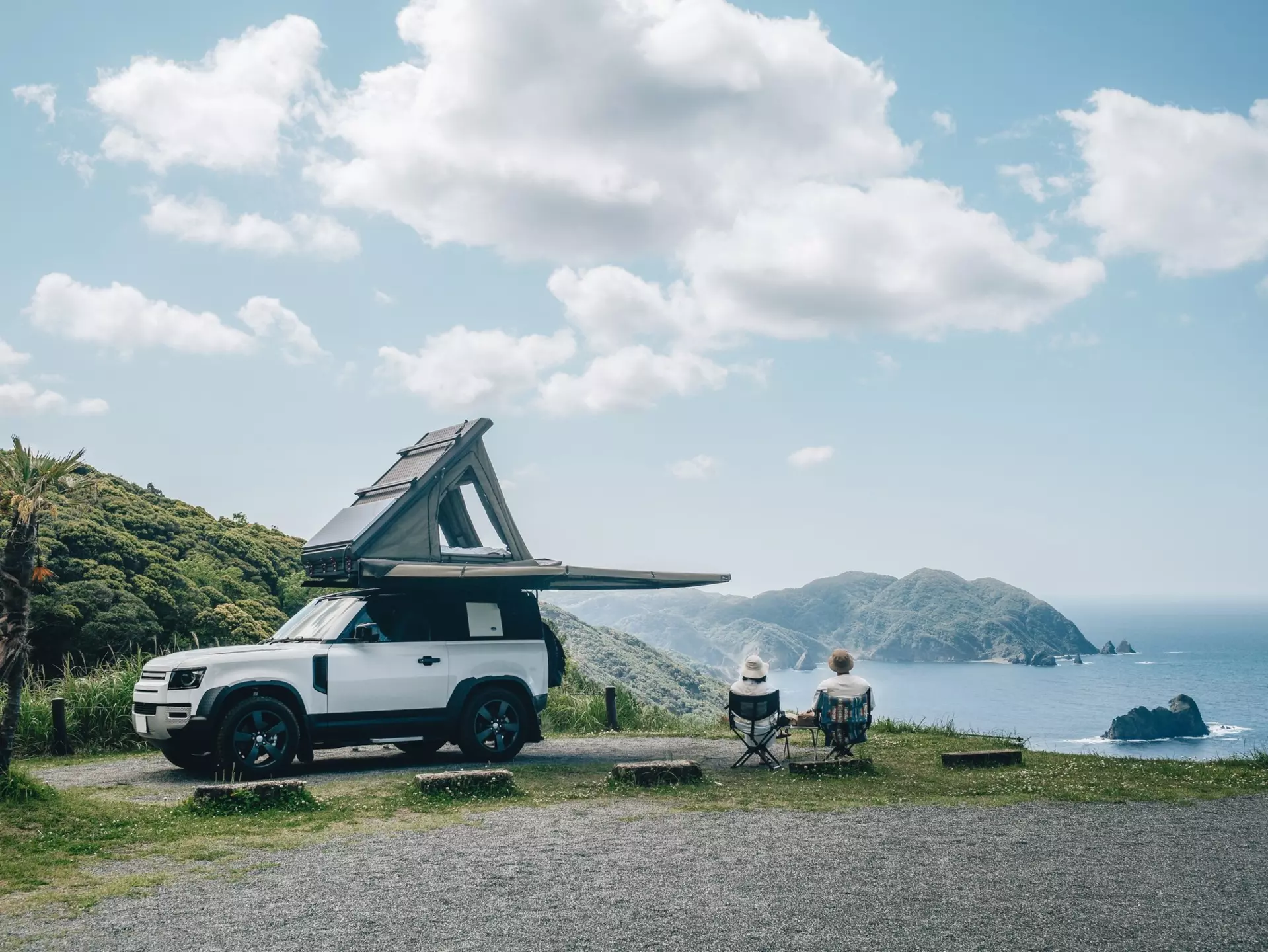 Couple sitting in a camping chair in nature with a view of Island in a beautiful blue sky with an overlanding car with a rooftop tent and an awning. Hokkaido 1, other: Kat Marsh