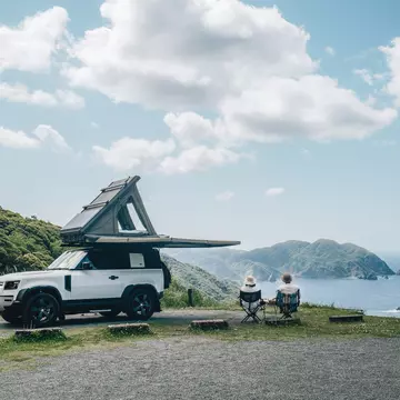 Couple sitting in a camping chair in nature with a view of Island in a beautiful blue sky with an overlanding car with a rooftop tent and an awning. Hokkaido 1, other: Kat Marsh