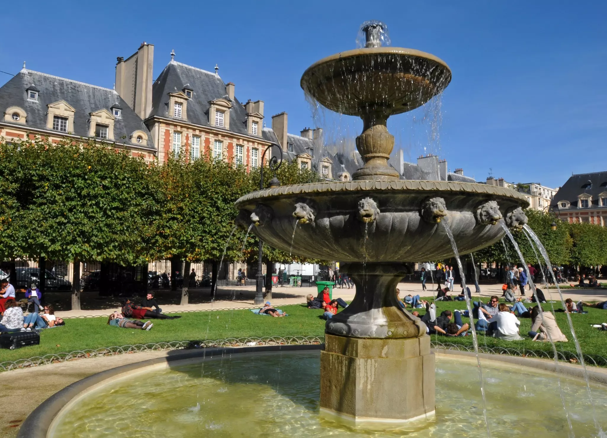 A fountain in a park with green grass where people are sitting.