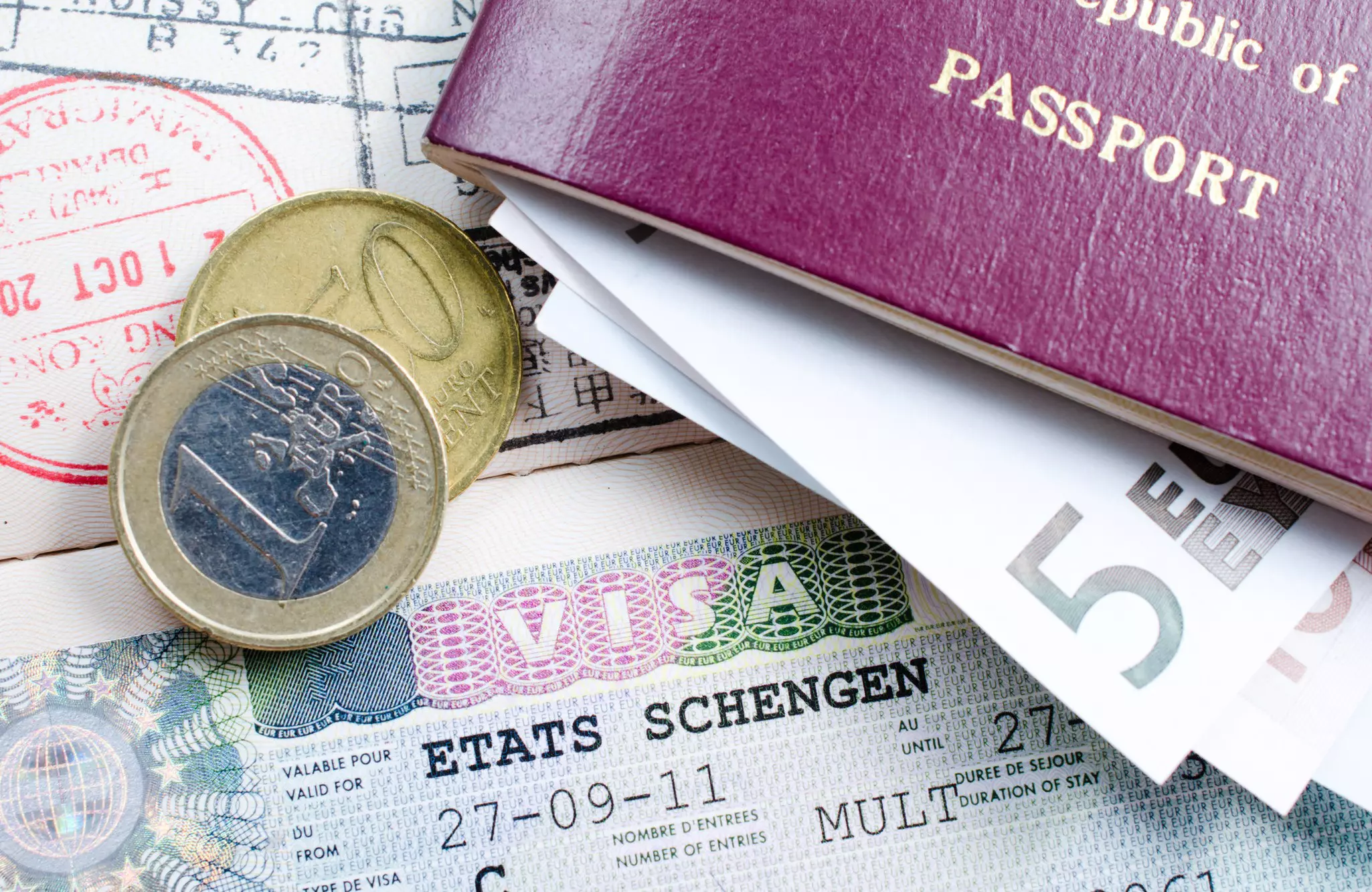 A pair of Euro coins sits on a Etats Schengen visa along with a maroon passport stuffed with paper Euro bills.