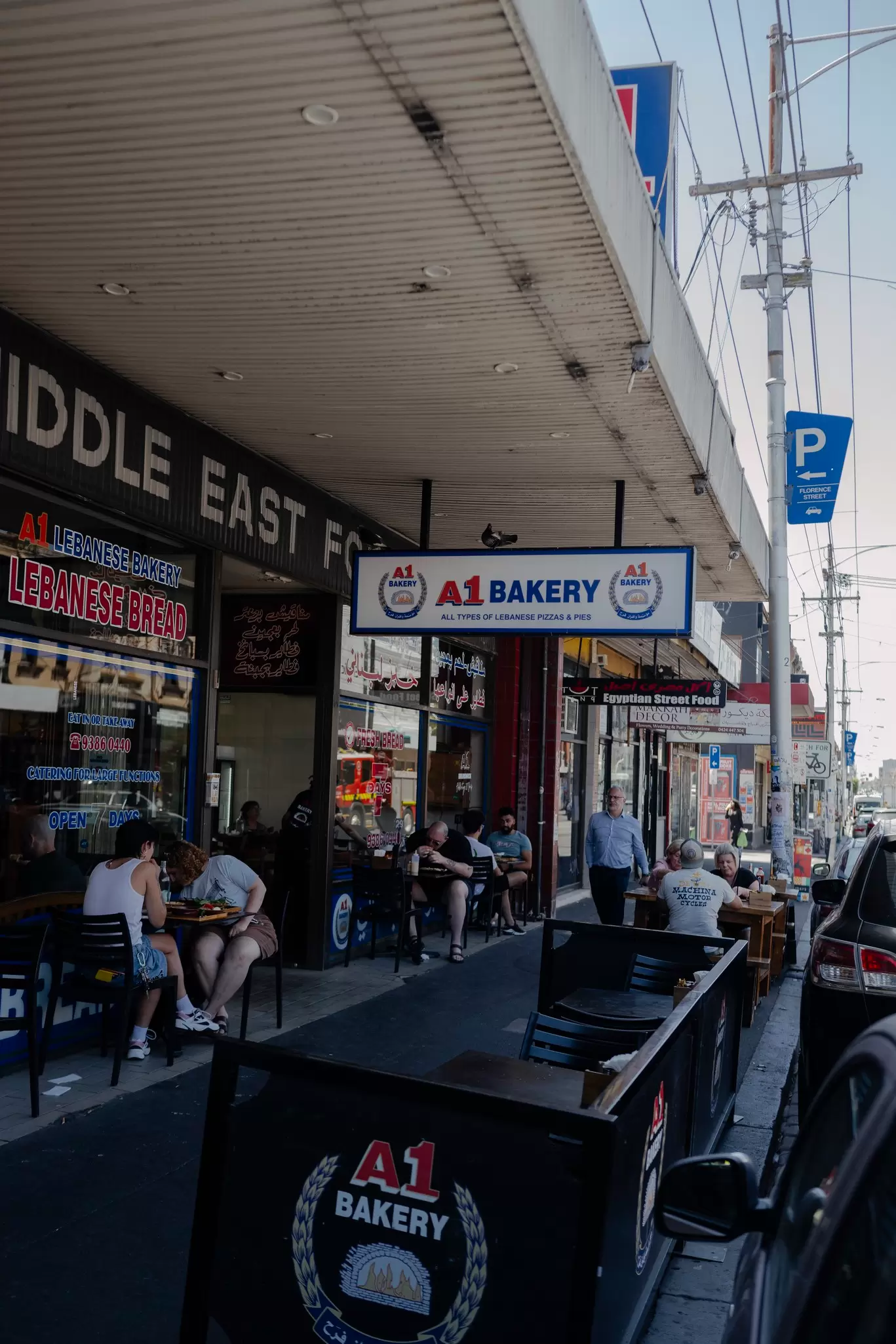 People eating at small tables outside a building with a sign that says "A1 Bakery" hanging in front in Melbourne, Australia.