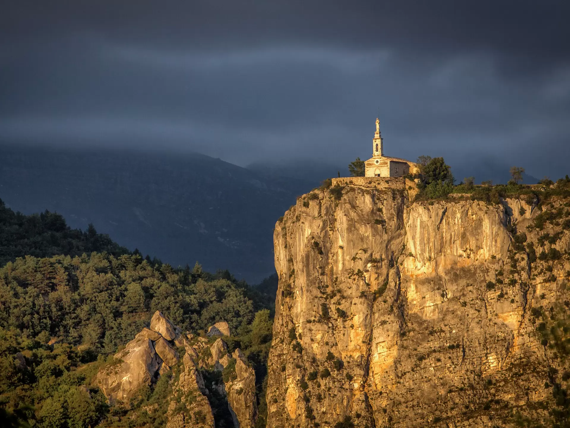 A chapel with a steeple at the top of a rocky cliff in warm sunlight.