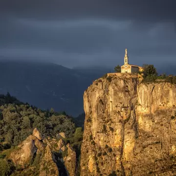 A chapel with a steeple at the top of a rocky cliff in warm sunlight.