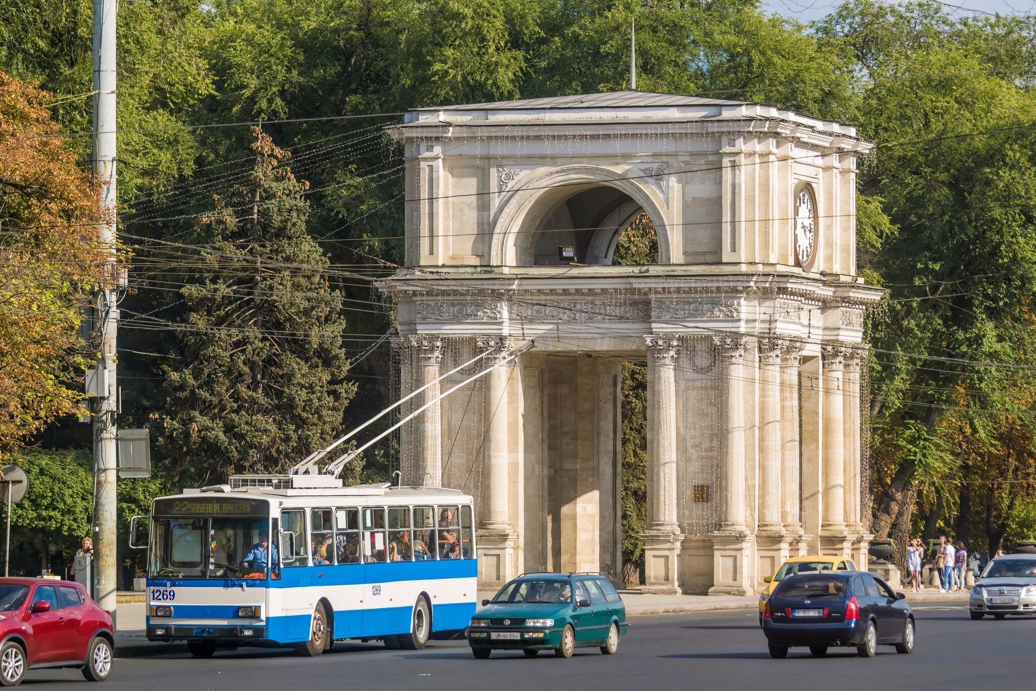 A stone triumphal arch with Corinthian columns and a clock face, trees behind and a blue-and-white trolleybus in front.
