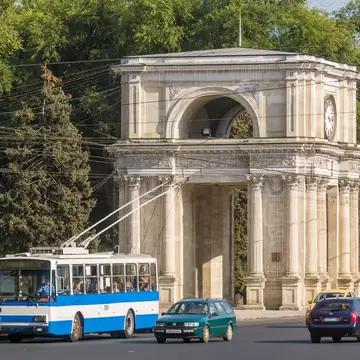 A stone triumphal arch with Corinthian columns and a clock face, trees behind and a blue-and-white trolleybus in front.