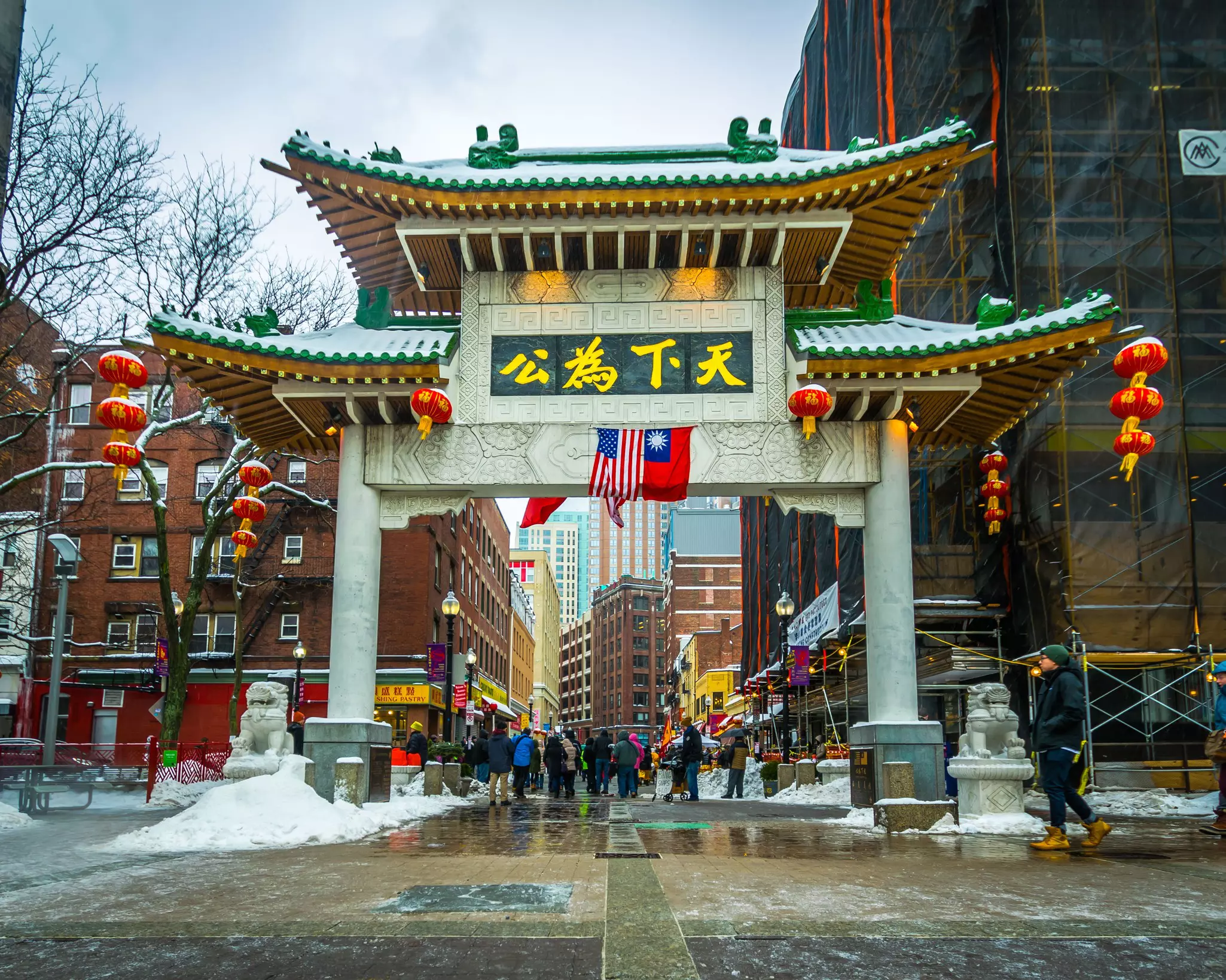A Chinese-style gateway to a neighborhood in a city. It's hung with red and gold lanterns. Large piles of snow line the sidewalks.